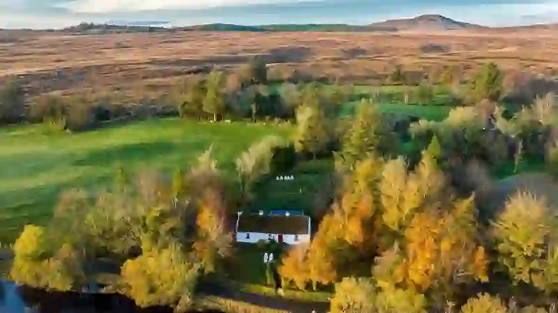 Aerial view of Keenaghan Cottage among autumn trees beside a lake, with rugged moorland and a hill beyond in County Fermanagh.