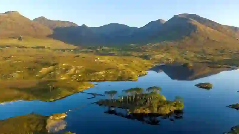 Lac Derryclare Lough dans le Connemara, dans le comté de Galway, avec petite île boisée, montagnes et reflets dans une eau calme.
