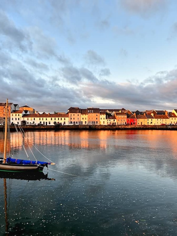Galway city's Long Walk and The Claddagh at sunset with colourful houses reflected in calm water.