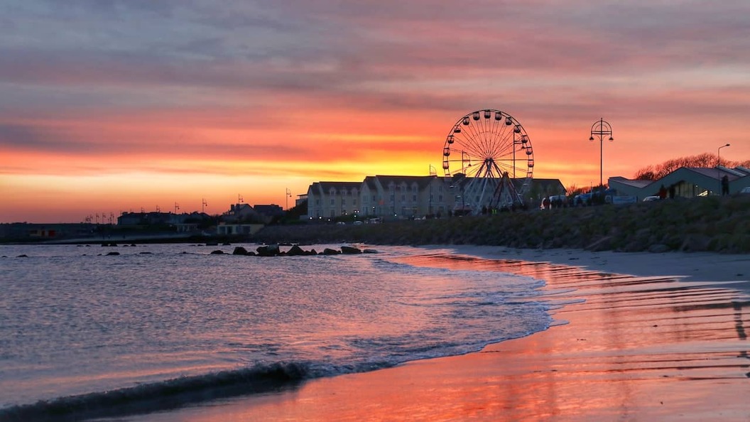 Promenade et plage de Salthill à Galway au soleil couchant, avec une grande roue se reflétant sur le rivage.