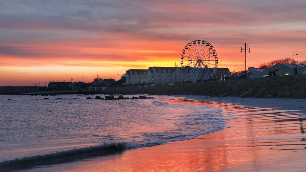 Ferris wheel beside Salthill beach at sunset with colourful skies reflected on the shoreline in Galway.