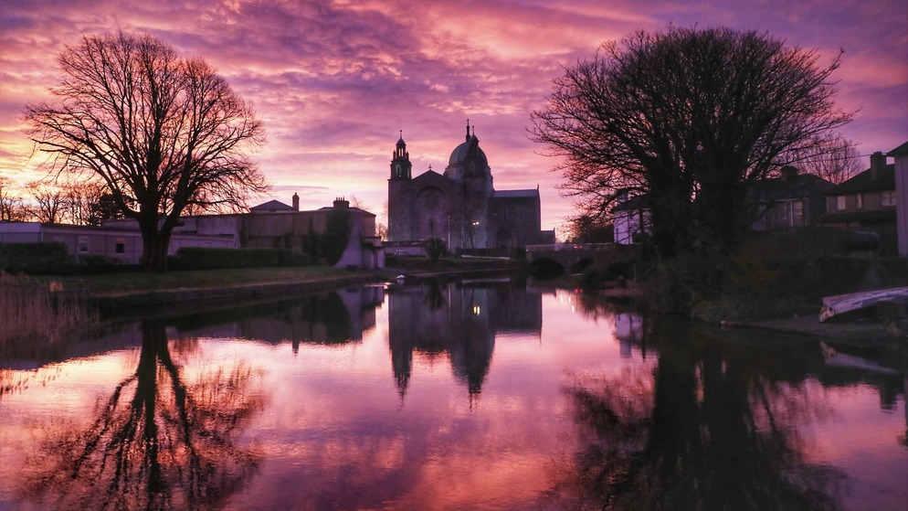 Galway Cathedral reflected in calm water beneath a purple sunset sky in Galway city.