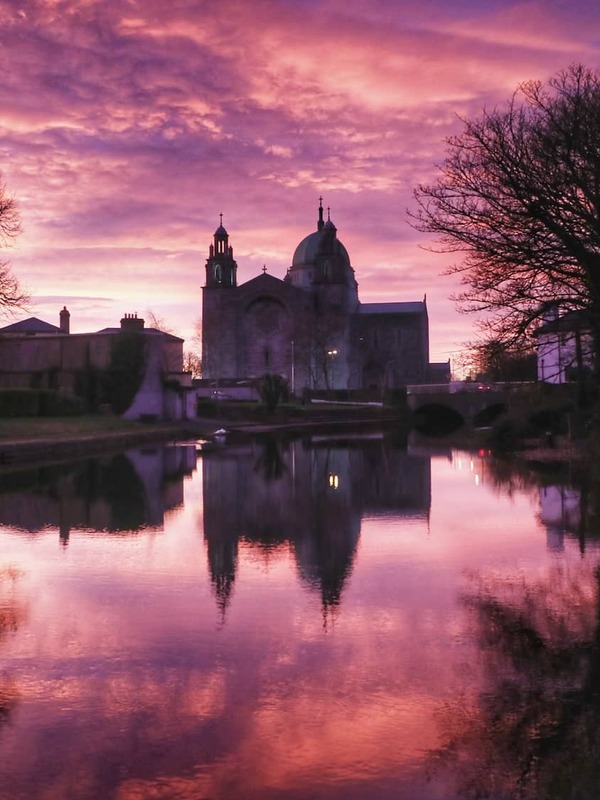 Galway Cathedral reflected in calm water beneath a purple sunset sky in Galway city.