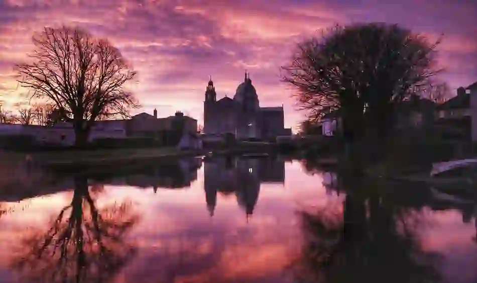 Galway Cathedral reflected in calm water beneath a purple sunset sky in Galway city.