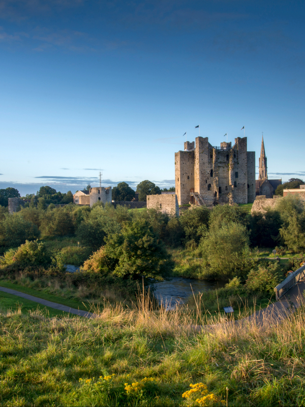Trim Castle rises above the River Boyne, with a footbridge, green parkland and blue evening sky in County Meath.