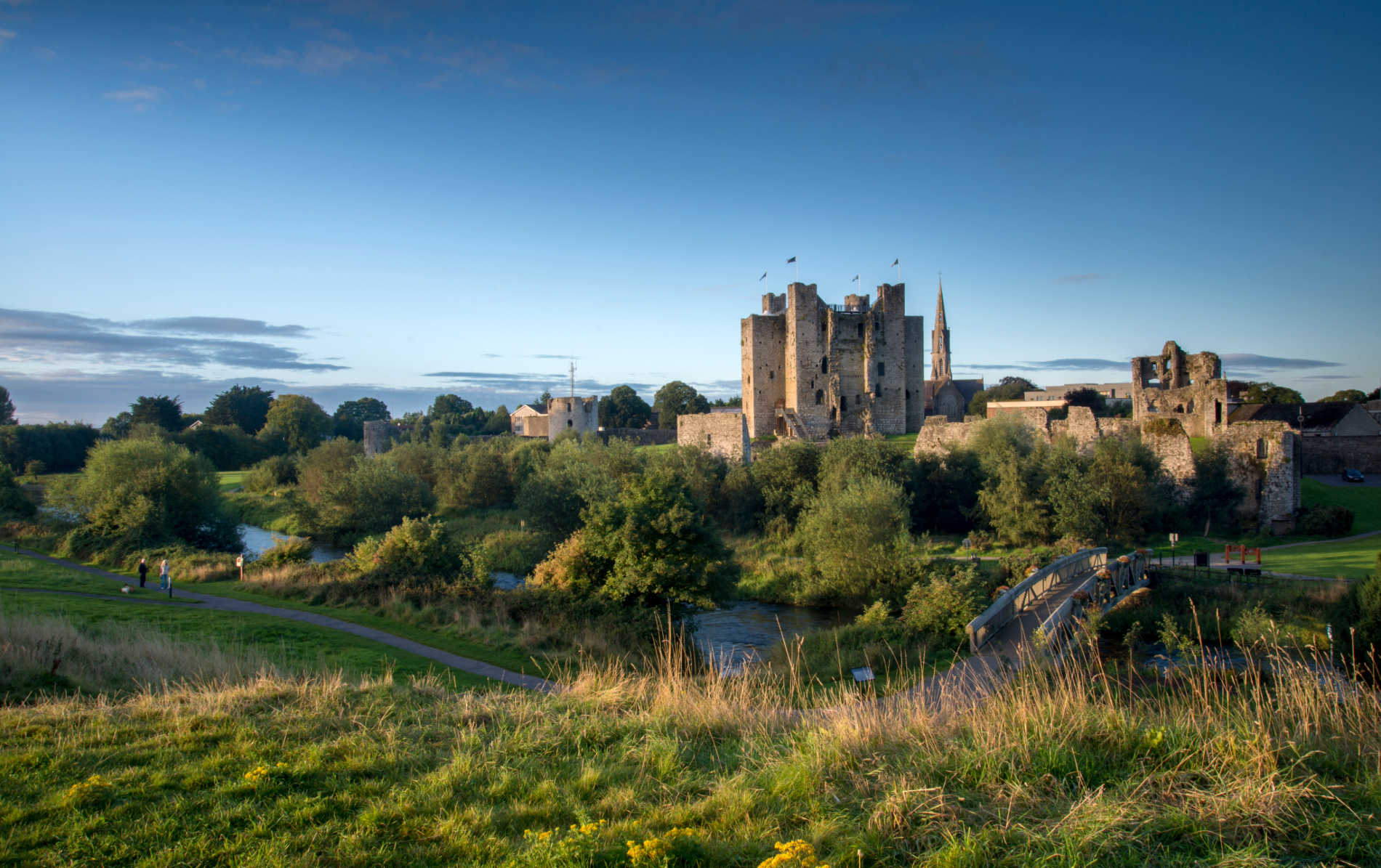 Trim Castle rises above the River Boyne, with a footbridge, green parkland and blue evening sky in County Meath.