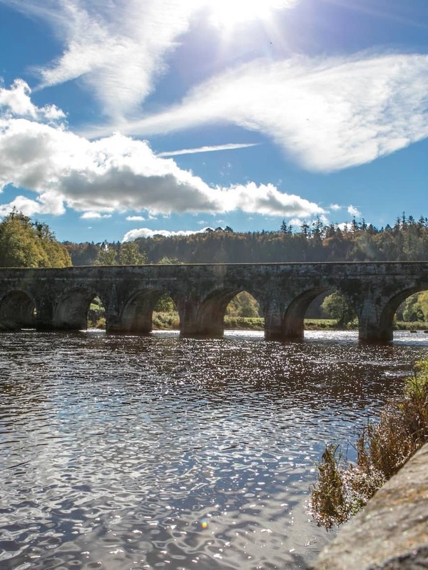 Stone arch bridge over the River Nore in Inistioge, County Kilkenny, with trees and blue sky reflected in the water.