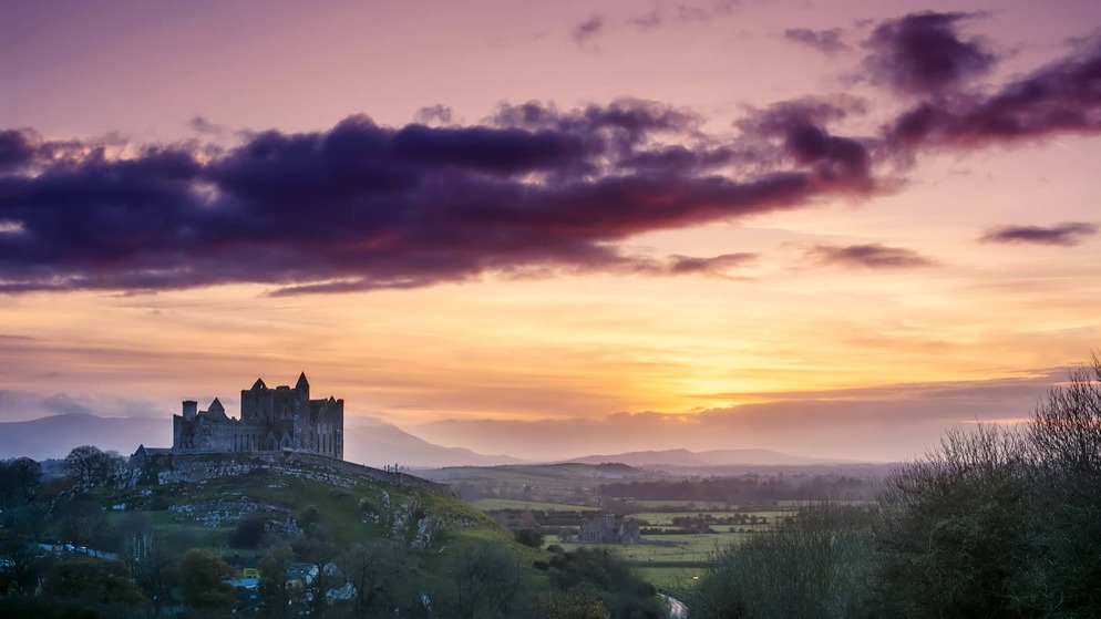 Rock of Cashel silhouetted at sunset over rolling countryside in County Tipperary, with vibrant pink and orange skies.
