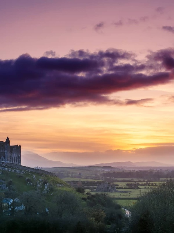 Rock of Cashel silhouetted at sunset over rolling countryside in County Tipperary, with vibrant pink and orange skies.