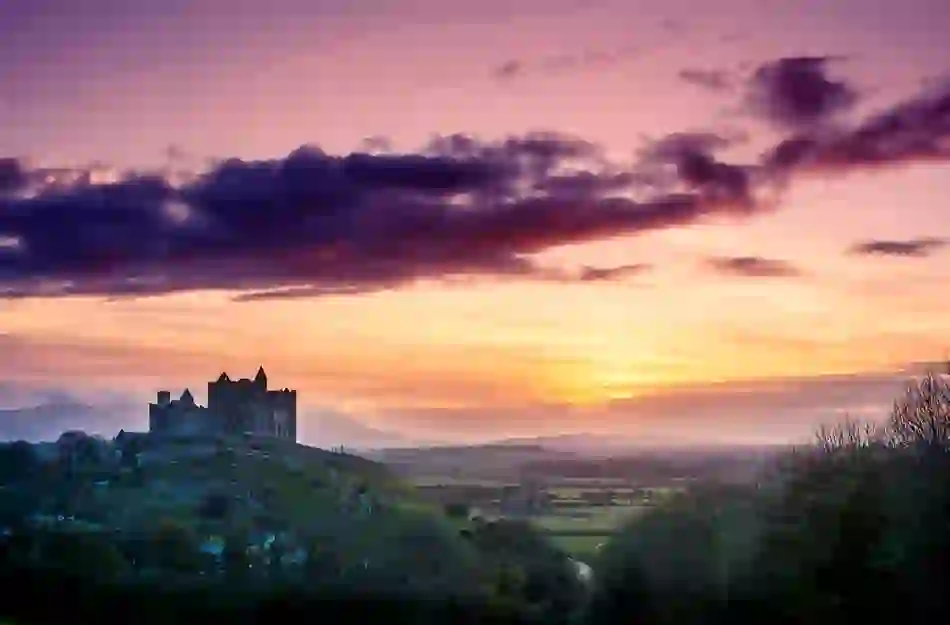 Rock of Cashel silhouetted at sunset over rolling countryside in County Tipperary, with vibrant pink and orange skies.