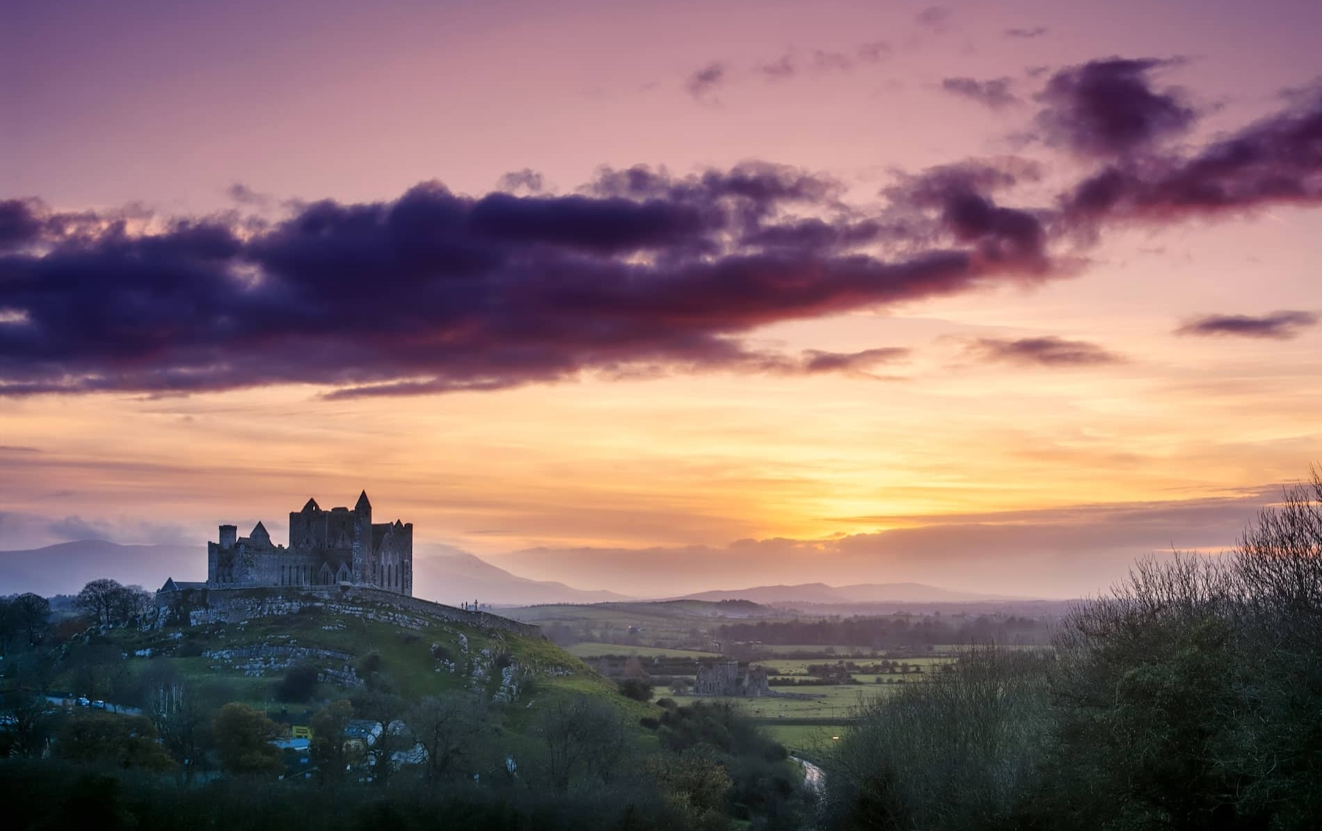 De Rock of Cashel bij zonsondergang boven het glooiende landschap van County Tipperary, met een kleurrijke roze en oranje lucht.