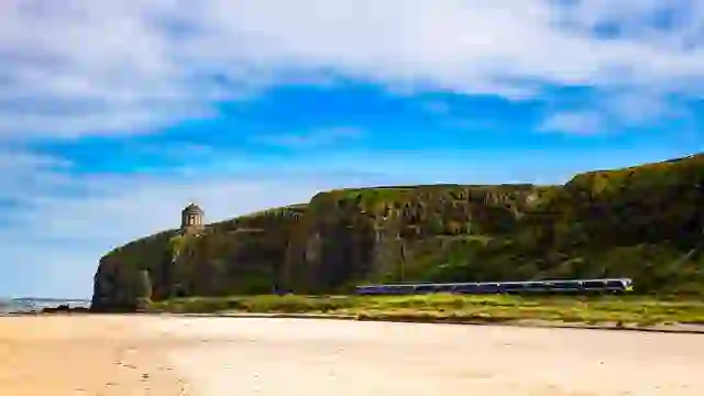 Der Mussenden-Tempel thront auf den imposanten Küstenklippen oberhalb von Downhill Strand, während unterhalb ein Zug entlang der Küste fährt.