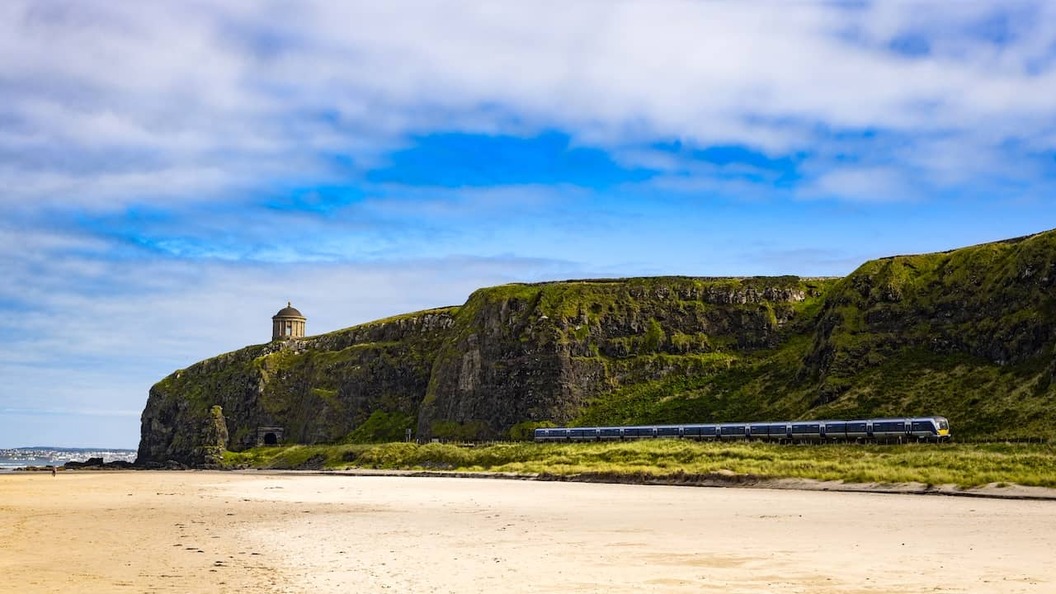 Der Mussenden-Tempel thront auf den imposanten Küstenklippen oberhalb von Downhill Strand, während unterhalb ein Zug entlang der Küste fährt.