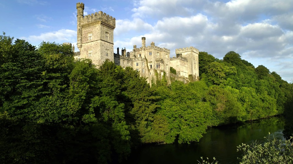 Lismore Castle, County Waterford, overlooking the River Blackwater, surrounded by lush green woodland under blue sky.