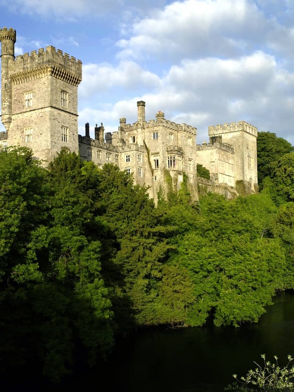 Lismore Castle, County Waterford, overlooking the River Blackwater, surrounded by lush green woodland under blue sky.