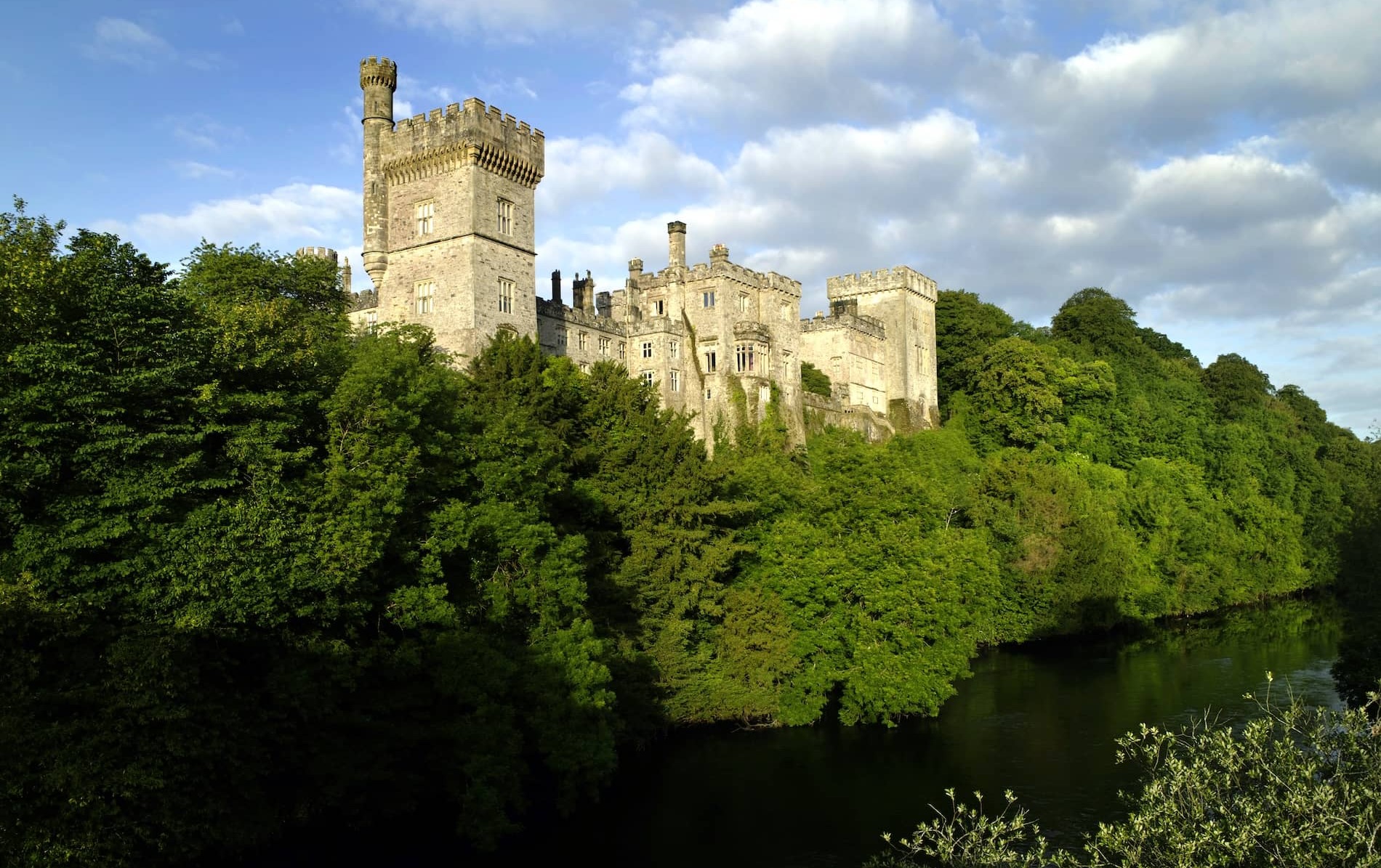 Lismore Castle, County Waterford, overlooking the River Blackwater, surrounded by lush green woodland under blue sky.