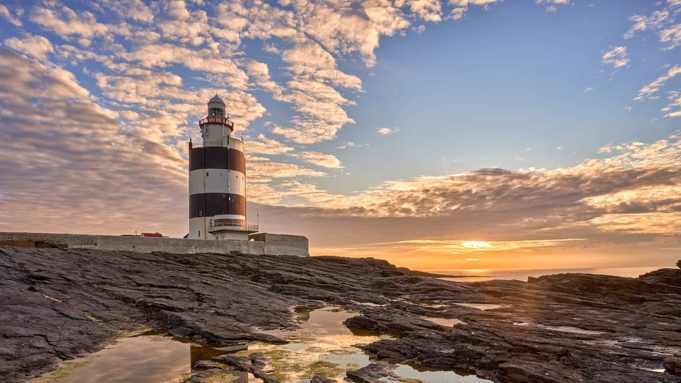 Hook Head Lighthouse in County Waterford reflected in rocky tidal pools at sunset beneath a dramatic sky.