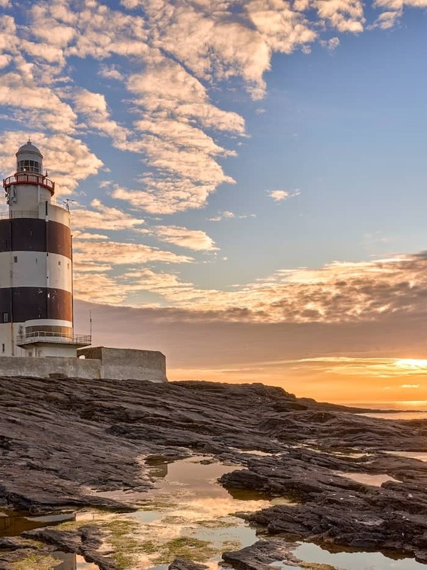 Hook Head Lighthouse in County Waterford reflected in rocky tidal pools at sunset beneath a dramatic sky.