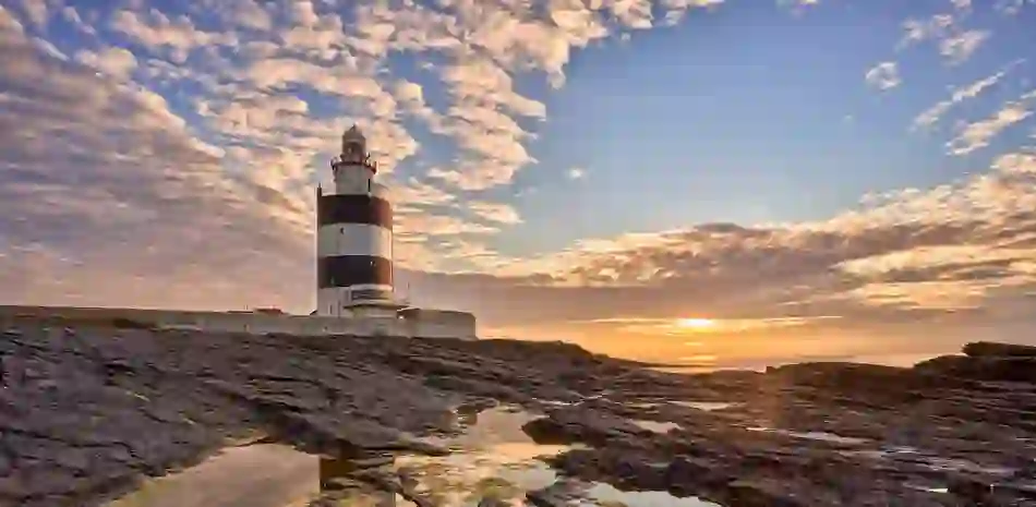 Hook Head Lighthouse in County Waterford reflected in rocky tidal pools at sunset beneath a dramatic sky.