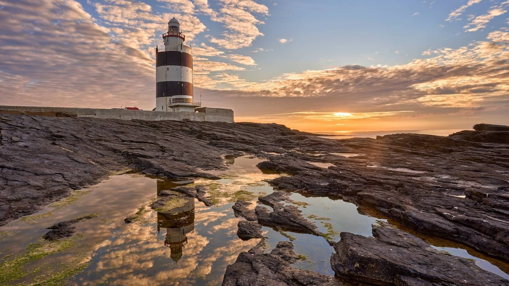 Hook Head Lighthouse in county Waterford weerspiegeld in rotsachtige branding bij zonsondergang onder een dramatische lucht.