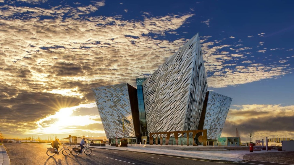 Titanic Belfast bij zonsondergang, met fietsers die langs de waterkant in het Titanic Quarter van Belfast rijden.