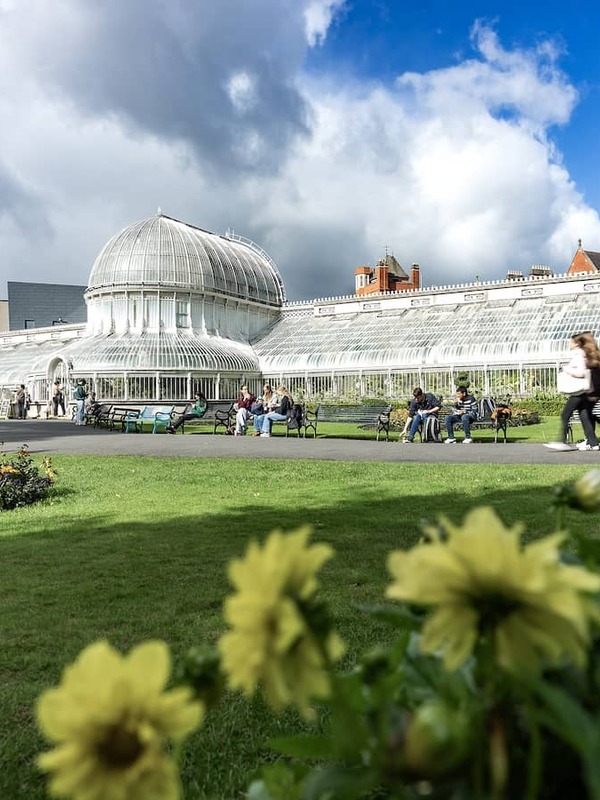 The Palm House at Belfast Botanic Gardens, with people relaxing on benches and flowerbeds in the foreground.