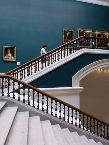 Visitor ascending the grand staircase in the National Gallery of Ireland beneath classic portraits.