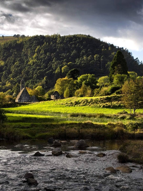 Het zonlicht breekt door de wolken boven de kloosterruïnes en de ronde toren van Glendalough, gelegen in een weelderige groene vallei bij een stromende beek.