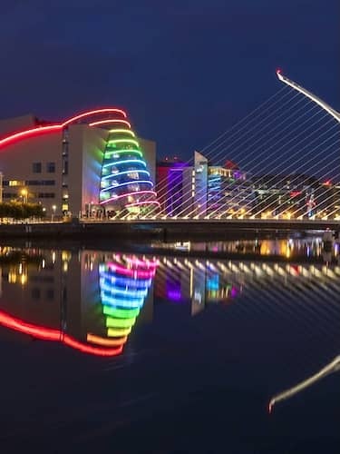 Night view of Samuel Beckett Bridge with Convention Centre lit in rainbow colours reflecting over the River Liffey.