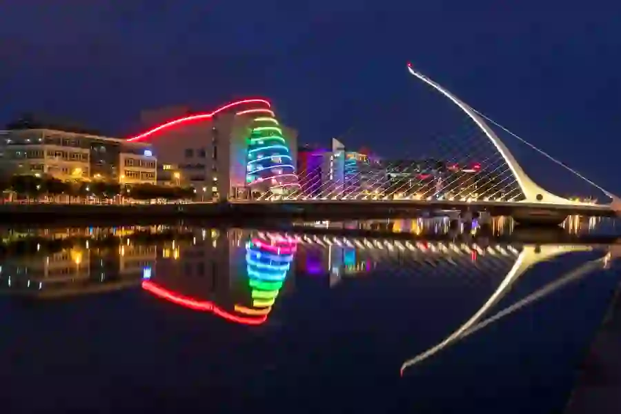 Colourfully lit Convention Centre and Samuel Beckett Bridge reflected in the River Liffey at night.