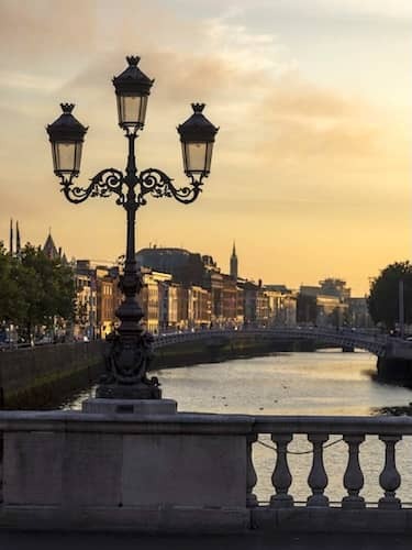 View of Ha’penny Bridge and River Liffey at sunset, with city buildings and church spire in central Dublin.