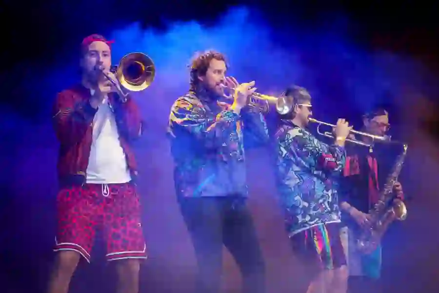 Musicians in vibrant outfits play brass instruments on stage, surrounded by dramatic purple and orange stage lighting.