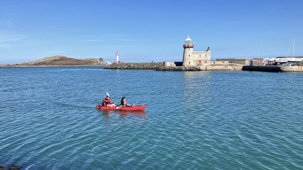 Two kayakers in red kayak on calm blue water near a historic lighthouse in Howth Harbour.
