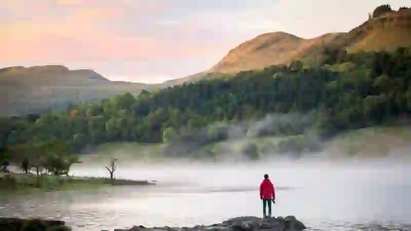 Person in a red coat standing by the misty shores of Glencar Lake in Leitrim, surrounded by forested hills at sunrise.