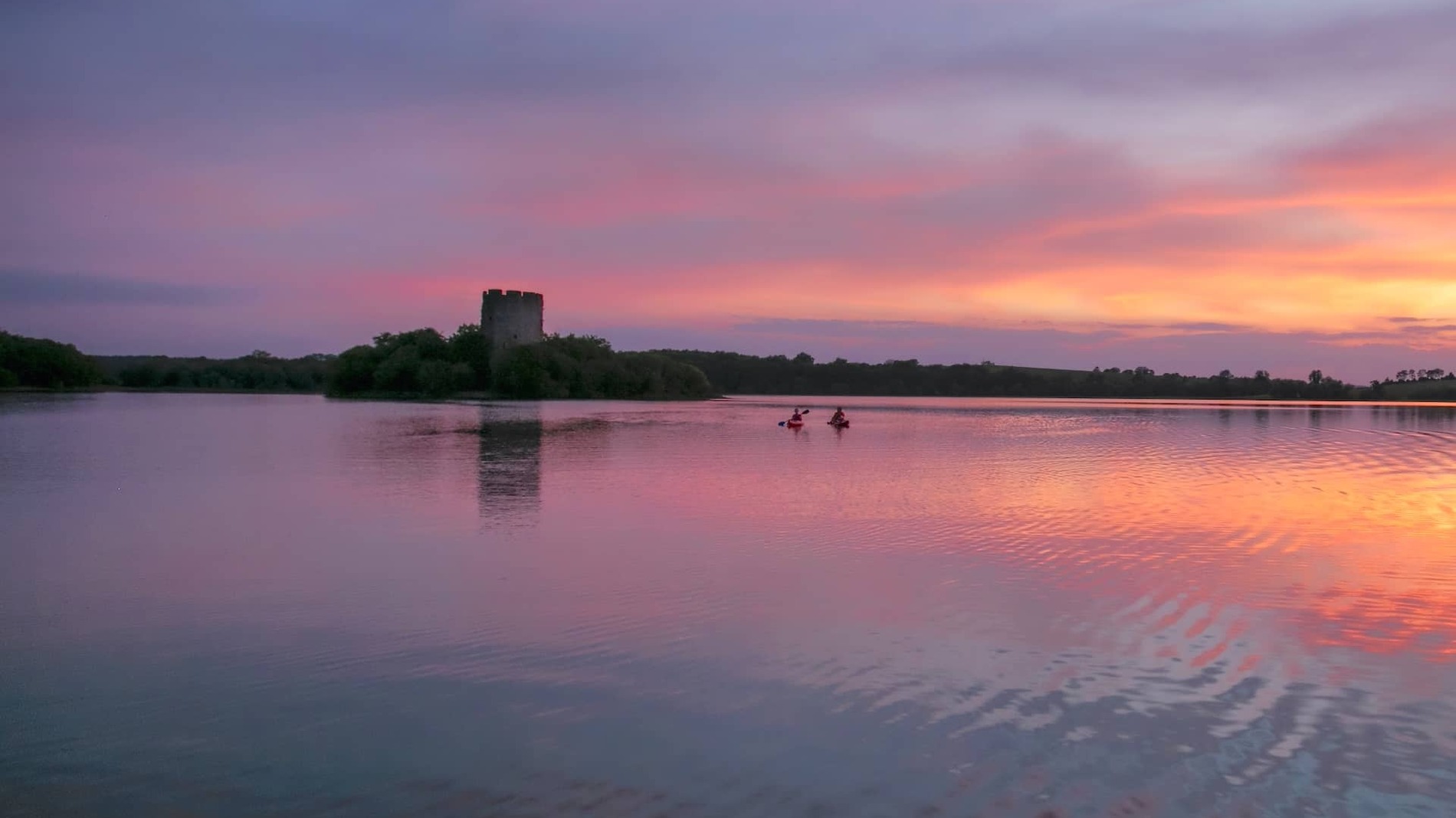 Two kayakers paddle across Lough Oughter in County Cavan at sunset, with a castle tower silhouetted against the pink sky.