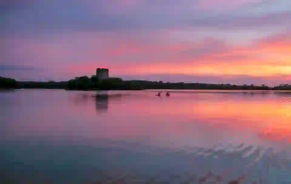 Two kayakers paddle across Lough Oughter in County Cavan at sunset, with a castle tower silhouetted against the pink sky.
