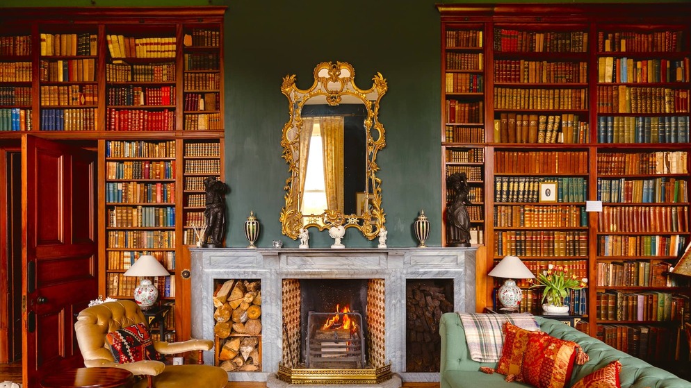 Elegant library at Clonalis House, County Roscommon, with tall wooden bookshelves, a marble fireplace, ornate mirror, and cosy fireside seating.