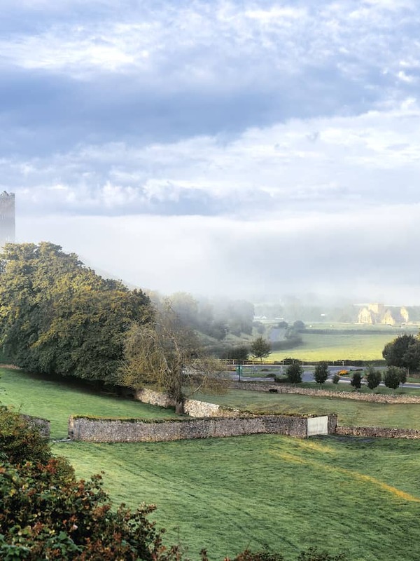Rock of Cashel rising above green farmland of County Tipperary, with medieval stone buildings framed by misty morning light.