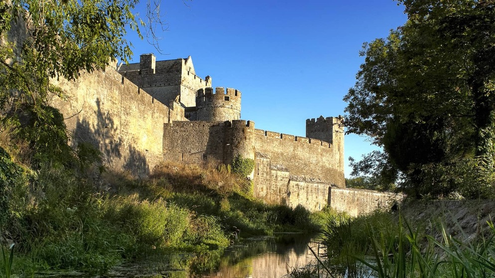 Cahir Castle reflected in a calm river, with stone walls and towers under a clear blue sky.