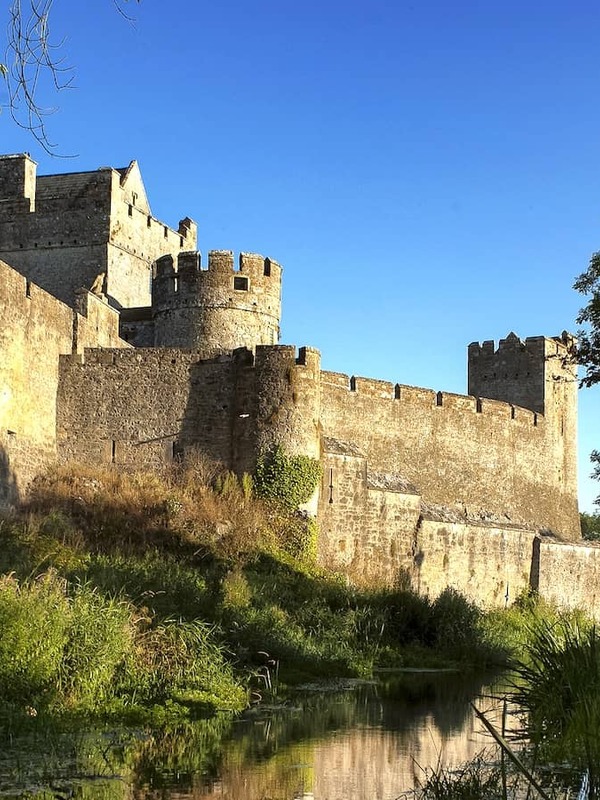 Cahir Castle reflected in a calm river, with stone walls and towers under a clear blue sky.
