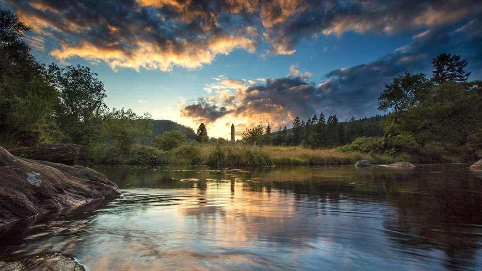 Sunset over the lakes at Glendalough, with the round tower framed by woodland and mountains.