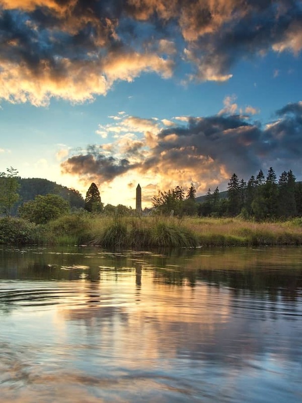 Sunset over the lakes at Glendalough, with the round tower framed by woodland and mountains.