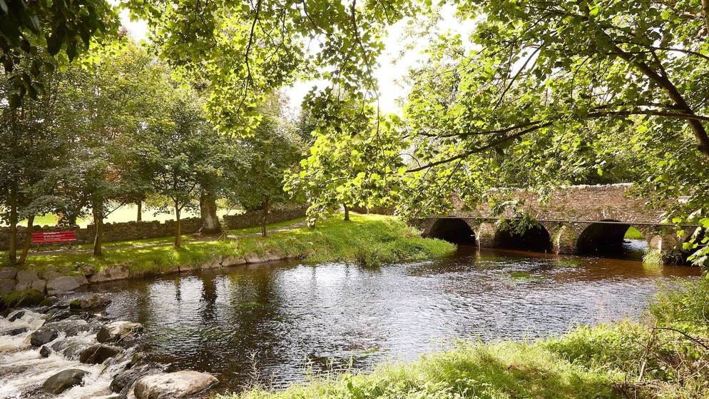 Sonnenbeschienener Ausblick auf den River Fane in Inniskeen, Grafschaft Monaghan, mit belaubten Bäumen und einer Steinbogenbrücke über ruhigem Wasser.
