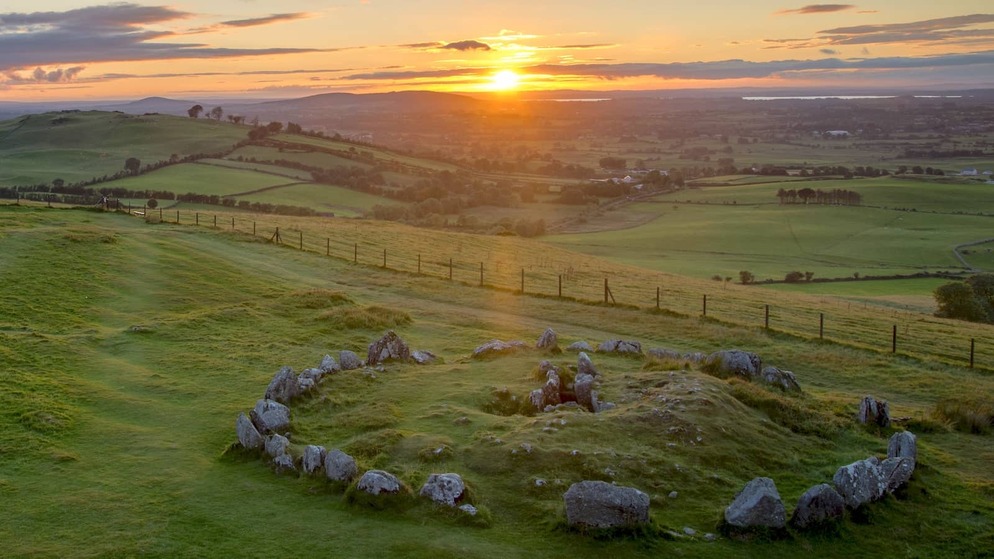 Sunset over the Loughcrew Cairns in County Meath, with a stone circle on grassy hills overlooking patchwork farmland.