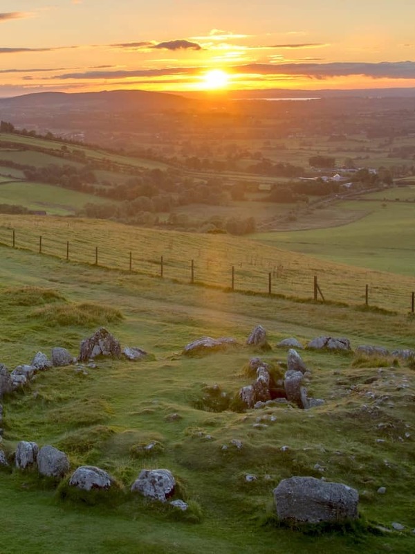 Tramonto sul Loughcrew Cairns nella contea di Meath, con un cerchio di pietre su verdi colline che si affaccia su un mosaico di terreni agricoli.
