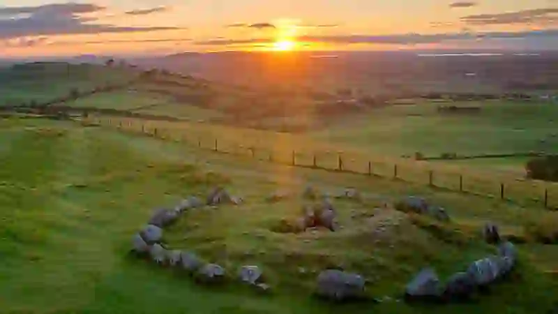 Tramonto sul Loughcrew Cairns nella contea di Meath, con un cerchio di pietre su verdi colline che si affaccia su un mosaico di terreni agricoli.