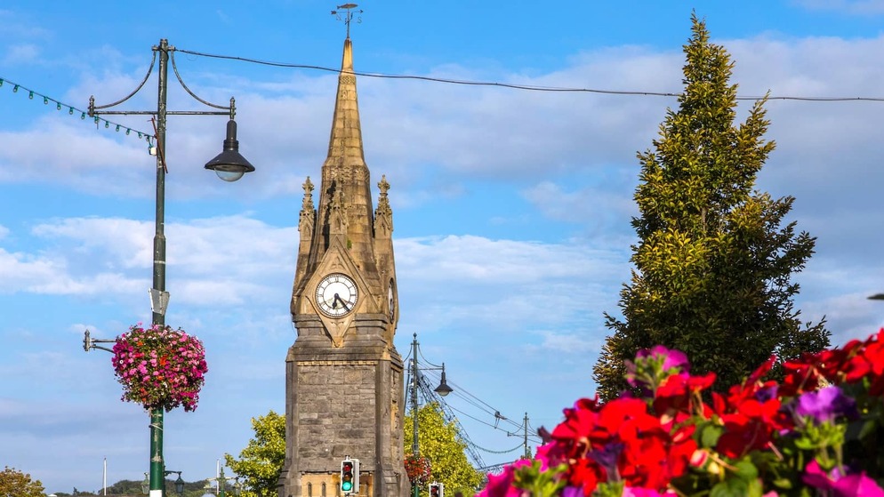 Clock tower rising above a city street in Waterford, framed by colourful flowers and hanging baskets under a bright blue sky.