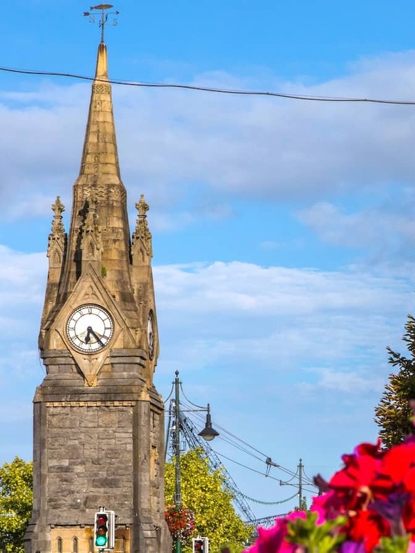 Clock tower rising above a city street in Waterford, framed by colourful flowers and hanging baskets under a bright blue sky.