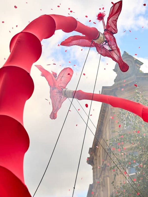 Red stilt-walking performers towering above a city street, with confetti falling during a festival.