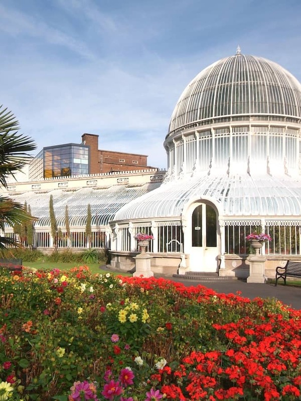 Glass-domed Palm House surrounded by vibrant summer flowerbeds and tropical plants in Belfast Botanic Gardens.